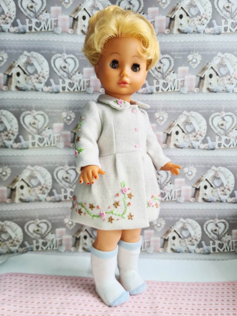 K&ouml;nnte beinhalten: Eine Vintage-Puppe mit blonden Haaren, die ein wei&szlig;es Kleid mit gestickten Blumen und blauen Socken tr&auml;gt. Die Puppe steht auf einer rosa-wei&szlig;en gemusterten Oberfl&auml;che.