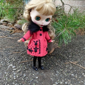 May include: A doll with blonde hair wearing a red coat with black embroidery and a black fur collar. The doll is wearing black tights and black shoes. The doll is standing on a gravel surface.