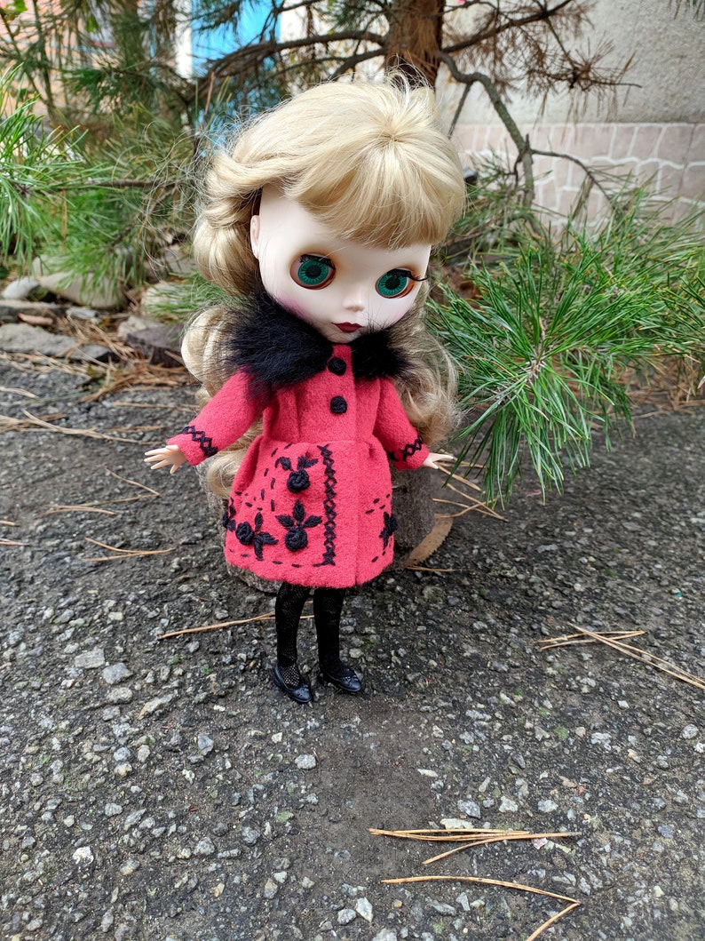 May include: A doll with blonde hair wearing a red coat with black embroidery and a black fur collar. The doll is wearing black tights and black shoes. The doll is standing on a gravel surface.