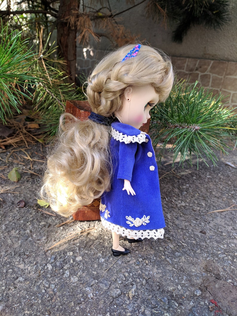 May include: A doll with long blonde hair styled in a braid, wearing a blue corduroy coat with white lace trim and floral embroidery. The doll is wearing black shoes and has a pink and blue hair clip.