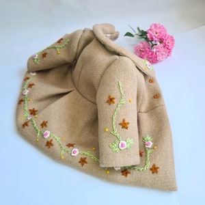 May include: A beige children's coat with embroidered flowers and vines in shades of green, pink, and brown.