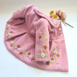 May include: A pink wool coat with embroidered floral details. The coat has a ruffled hem and long sleeves with a vine and flower design.