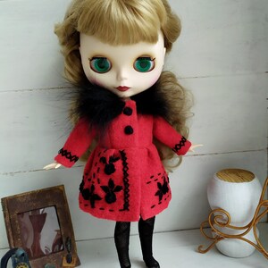 May include: A red felt doll coat with black faux fur trim and black embroidered flowers. The doll has blonde hair, green eyes, and is wearing black tights and black shoes.