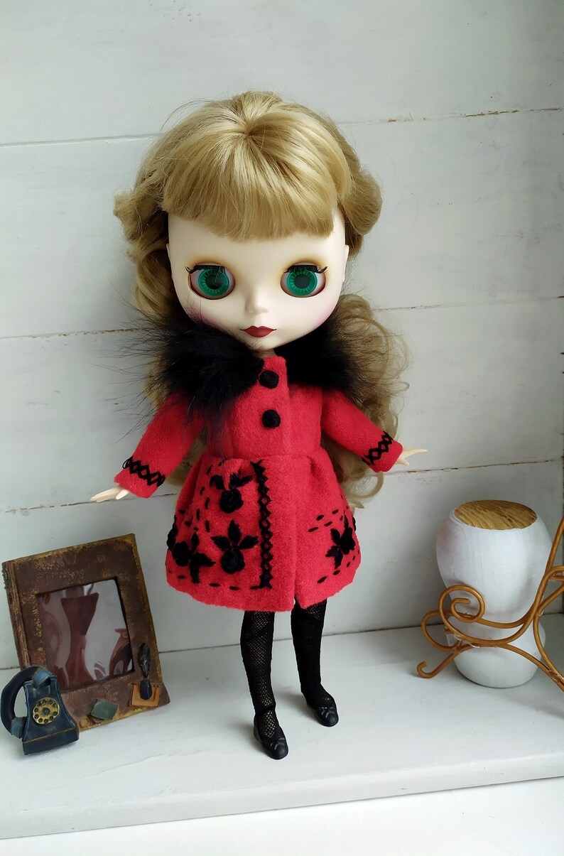 May include: A red felt doll coat with black faux fur trim and black embroidered flowers. The doll has blonde hair, green eyes, and is wearing black tights and black shoes.