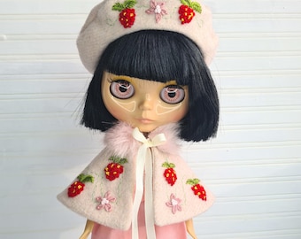 May include: A pink doll with black hair wearing a pink beret with embroidered strawberries and a pink cape with embroidered strawberries and flowers. The doll is wearing a pink dress and a white ribbon.