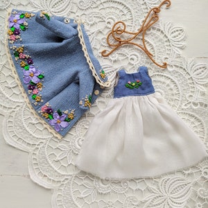 以下が含まれることがあります： デニムジャケットと白い花の刺繍のドレス。 ジャケットは、紫、ピンク、黄色の花のアクセントが付いた水色のデニム生地です。 ドレスは白いスカートと花の刺繍が施された青いボディスです。 金色のハンガーも付いています。