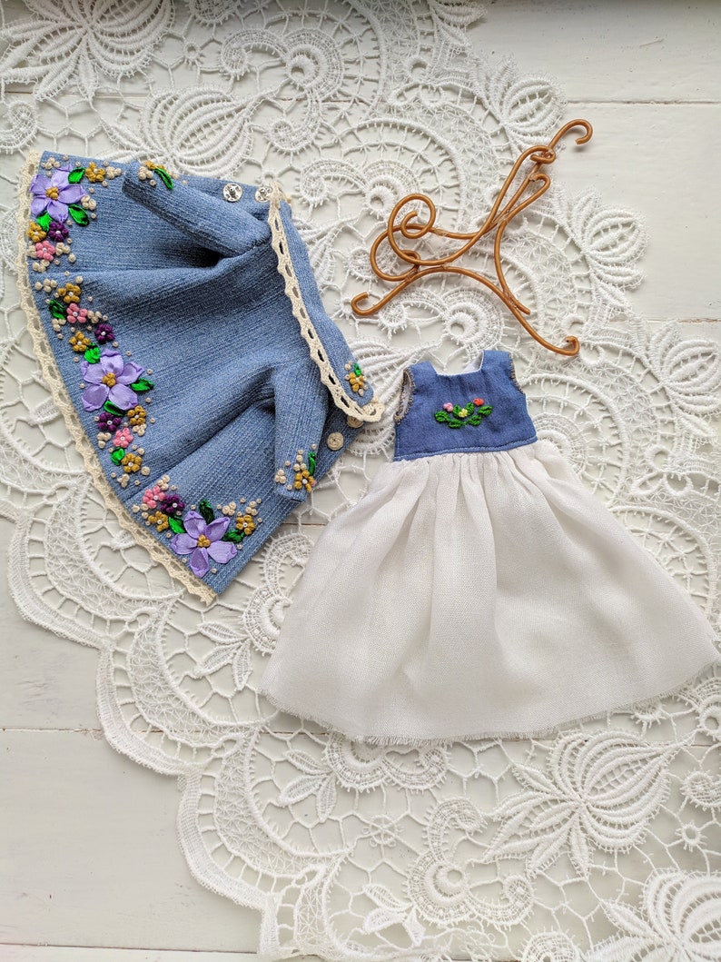 以下が含まれることがあります： デニムジャケットと白い花の刺繍のドレス。 ジャケットは、紫、ピンク、黄色の花のアクセントが付いた水色のデニム生地です。 ドレスは白いスカートと花の刺繍が施された青いボディスです。 金色のハンガーも付いています。
