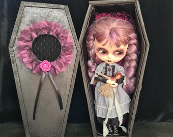 May include: A pink and white doll with purple hair is lying in a gray coffin with a pink and black floral design. The coffin is open and the doll is holding a small doll.