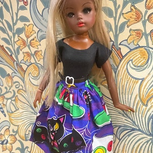 May include: A doll with long blonde hair and brown skin wearing a black top and a purple skirt with a black cat, spider web, and eye print.