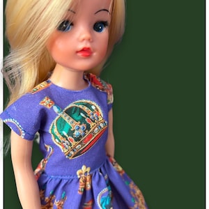 Può includere: Una bambola con i capelli biondi che indossa un vestito viola con un motivo a corona. Il vestito ha maniche corte e una gonna ampia.