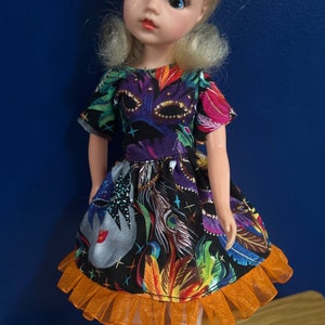 Könnte beinhalten: Eine Puppe, die ein farbenfrohes Kleid mit einem Maskenmuster und einem orangefarbenen Rüschenbesatz trägt. Die Puppe hat blonde Haare und blaue Augen.
