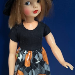 May include: A doll wearing a black witch hat and a black and orange dress with a pumpkin pattern. The doll has blonde hair and blue eyes.