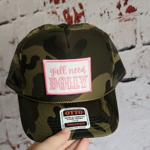 May include: A camouflage trucker hat with a pink patch that says "y'all need DOLLY". The hat is made by OTTO and has a foam front.