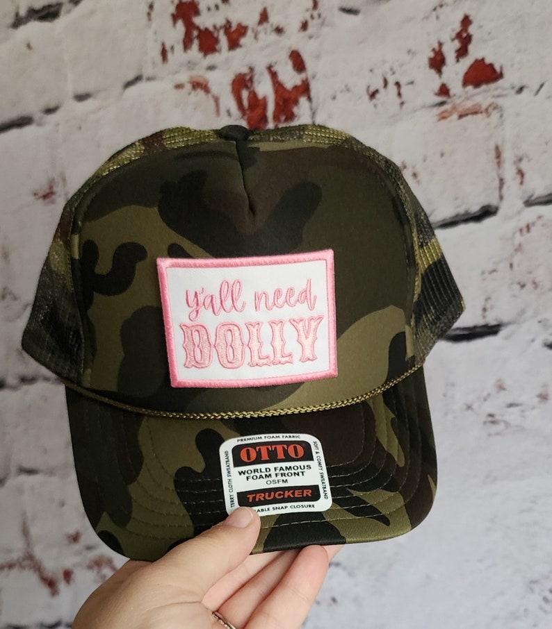 May include: A camouflage trucker hat with a pink patch that says "y'all need DOLLY". The hat is made by OTTO and has a foam front.