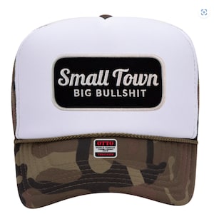 May include: A white trucker hat with a black rectangle patch that reads "Small Town Big Bullshit" in white script. The brim and lower portion of the hat feature a camouflage pattern. The hat has a brown top and a white mesh back.