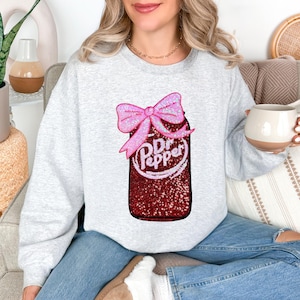 May include: A light gray sweatshirt featuring a sequined Dr. Pepper design with a pink bow. The Dr. Pepper logo is in white script. The sweatshirt is a casual style, suitable for everyday wear.