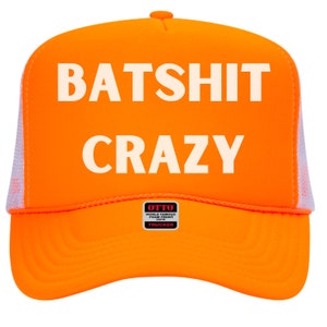 May include: Bright orange trucker hat with the words "BATSHIT CRAZY" in large, white capital letters. The hat has a white mesh back and a curved brim. The Otto brand logo is visible on the front.