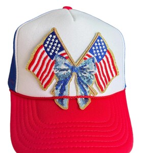 May include: A red, white, and blue trucker hat featuring a central patch of two crossed American flags tied together with a shimmering light blue sequined bow. The hat has a solid red brim, a white front panel, and a blue mesh back with a red cord.
