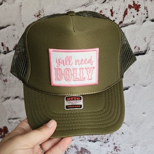 May include: A green trucker hat with a pink patch that says "y'all need DOLLY". The hat has a black mesh back and a white label that says "OTTO WORLD FAMOUS FOAM FRONT 05FM TRUCKER".