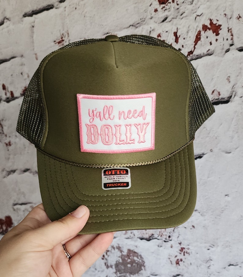 May include: A green trucker hat with a pink patch that says "y'all need DOLLY". The hat has a black mesh back and a white label that says "OTTO WORLD FAMOUS FOAM FRONT 05FM TRUCKER".