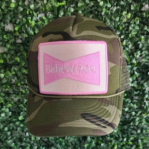 May include: Camouflage trucker hat with a pink and white patch that says "Babeweiser" in white text.
