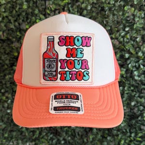 May include: A white and pink trucker hat with a colorful patch that says "Show Me Your Titos" and a bottle of Tito's Vodka. The hat has a black mesh back and a white front.