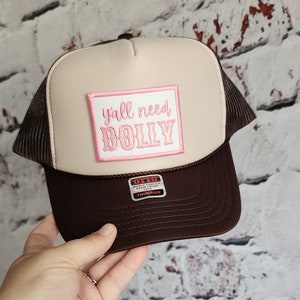 May include: A brown and beige trucker hat with a pink patch that says "y'all need dolly" on the front. The hat has a brown mesh back and a black label that says "Otto World Famous Foam Front OS/FM Trucker".