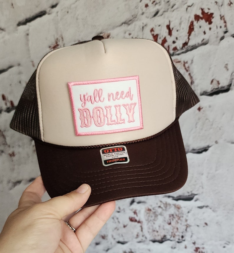 May include: A brown and beige trucker hat with a pink patch that says "y'all need dolly" on the front. The hat has a brown mesh back and a black label that says "Otto World Famous Foam Front OS/FM Trucker".