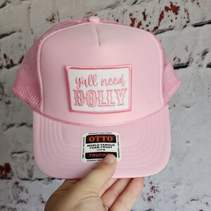 May include: Pink trucker hat with a white patch that says "y'all need DOLLY" in pink lettering. The hat has a mesh back and a black and red label that says "OTTO WORLD FAMOUS FOAM FRONT OSFM TRUCKER DURABLE SNAPBACK".