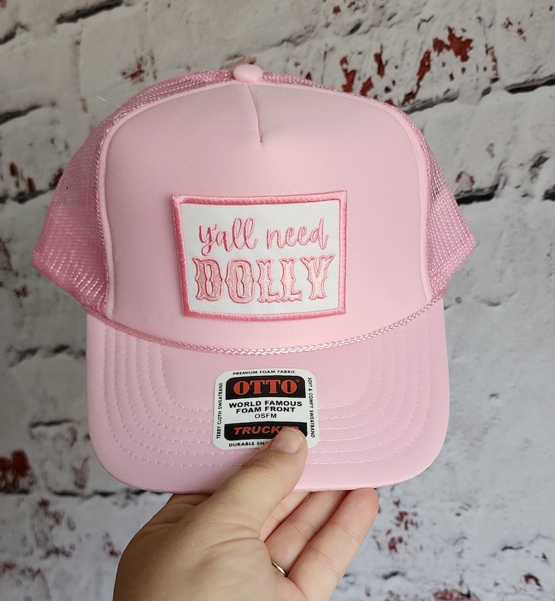 May include: Pink trucker hat with a white patch that says "y'all need DOLLY" in pink lettering. The hat has a mesh back and a black and red label that says "OTTO WORLD FAMOUS FOAM FRONT OSFM TRUCKER DURABLE SNAPBACK".