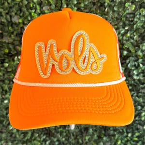 May include: Orange trucker hat with a white brim and a white mesh back. The hat has a large, gold-colored patch with the word "Yola" in rhinestones.