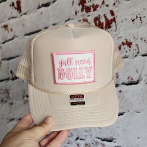 May include: A beige trucker hat with a pink and white patch that says "y'all need DOLLY". The hat has a mesh back and a black and red label that says "OTTO WORLD FAMOUS TRUCKER CAP"