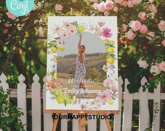 Könnte beinhalten: Ein weißes Willkommensschild auf einer Holzstaffelei, mit einem Foto einer Person in einem Blumenkleid. Das Schild ist mit einem Blumen- und Zitronenrand verziert, mit dem Text "Welcome Emily Johnson". Das Schild steht vor einem weißen Zaun und rosa Rosen.