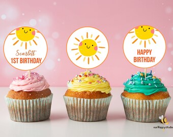 May include: Three cupcakes with yellow, pink, and green frosting. Each cupcake has a round, white paper topper with a smiling sun design. The toppers have the text "Scarlett 1st Birthday" and "Happy Birthday".