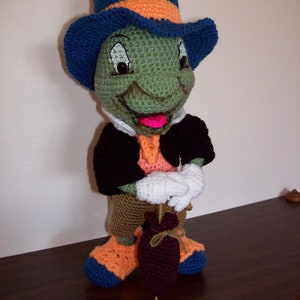 Puede incluir: Un títere de Jiminy Cricket de crochet verde y marrón que lleva un sombrero de copa azul y naranja y una chaqueta negra. El títere sostiene una bolsa marrón y morada.