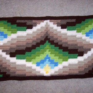 Op de afbeelding: Een gehaakt kussen met een geometrisch patroon in tinten bruin, groen, wit, blauw en geel.