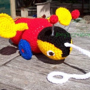 May include: A crocheted bee toy with yellow and red wings, a black and yellow body, and a white pull string. The bee has blue wheels and is designed by Joey's Creations NZ.