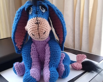 Pode incluir: Um burro de peluche de crochê feito com fio azul e roxo. O burro tem um nariz rosa, olhos pretos e uma cauda preta com um laço rosa.