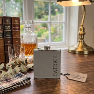 Peut inclure: Une flasque en similicuir gris avec un bouchon et une base en métal argenté, portant le nom PATRICK gravé verticalement sur le devant. Une petite étiquette rectangulaire beige attachée par un cordon noir indique THE GROOMSMEN COLLECTION.