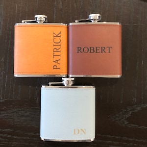 May include: Three personalised leather-wrapped hip flasks. The flasks are brown, orange, and light blue. The names "Patrick", "Robert", and "DN" are engraved on the flasks.