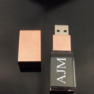 May include: A rose gold USB flash drive with a clear acrylic case. The drive is engraved with the name "AJM".