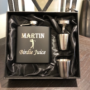 May include: A black flask engraved with the name "MARTIN" and the text "Birdie Juice". The flask is presented in a black box with a black satin lining. The box also includes a silver funnel and two silver shot glasses.