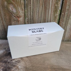 May include: A white box with a label that says "Mystery Slabs" and "5 slabs for cabbing and lapidary projects". The label also includes the text "Rock and Road Rockhounding" and the website address "www.etsy.com/shop/RockandRoad".