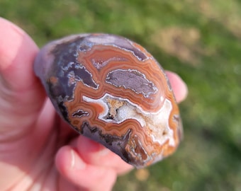 May include: A close-up of a brown, orange, and white agate stone with intricate banding patterns. The stone is smooth and polished, and it has a natural, organic shape.