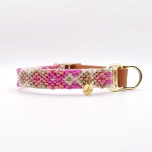 Könnte beinhalten: Ein gewebtes Haustierhalsband mit einem geometrischen Muster in Rosa-, Weiß- und Beigetönen. Das Halsband verfügt über einen beigefarbenen Lederriemen, goldfarbene Metallbeschläge und eine kleine runde goldfarbene Metallplakette mit der Aufschrift ALEIGA.