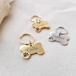 Könnte beinhalten: Drei knochenförmige Haustier-ID-Tags. Zwei sind goldfarben und einer silberfarben. Die goldenen Tags tragen die Namen "Calla" und "Mayla" eingraviert, mit Pfotenabdrücken. Das silberne Tag ist mit "Barney" graviert. Alle haben einen kleinen Ring zur Befestigung.