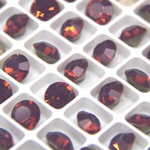 May include: A tray filled with numerous faceted, dark red-purple gemstones. The gems are arranged in a grid pattern within a white plastic organizer. Various shapes are visible, including round, oval, and teardrop.