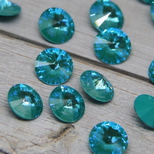 Pu&ograve; includere: Una collezione di strass ovali e rotondi, sfaccettati, color teal iridescente. Le pietre preziose sono sparse su una superficie di legno grigio chiaro. Le gemme hanno una lucentezza brillante, adatte per la creazione di gioielli o progetti artigianali.
