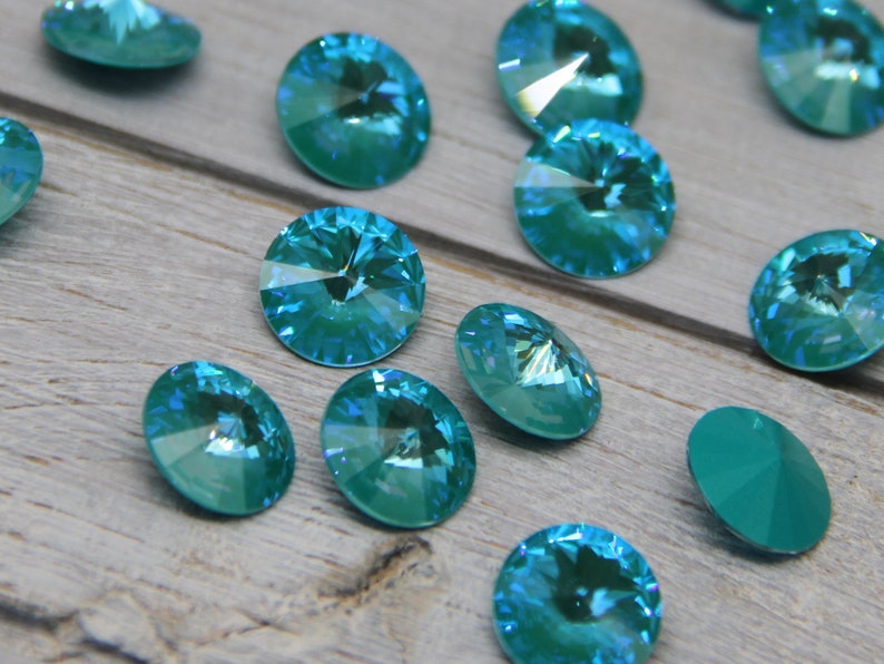 Pu&ograve; includere: Una collezione di strass ovali e rotondi, sfaccettati, color teal iridescente. Le pietre preziose sono sparse su una superficie di legno grigio chiaro. Le gemme hanno una lucentezza brillante, adatte per la creazione di gioielli o progetti artigianali.