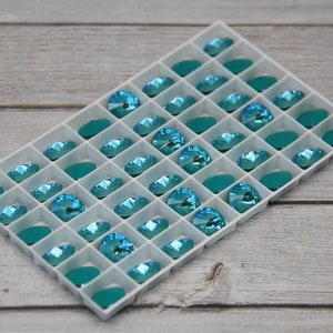 Pu&ograve; includere: Un vassoio bianco pieno di strass color acquamarina e pietre ovali color teal. Il vassoio &egrave; posto su uno sfondo chiaro con venature del legno, creando un contrasto tra le gemme luminose e lo sfondo neutro.